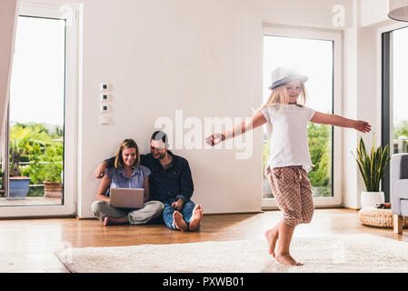 Little girl with hat having fun at home, parents using laptop in background Stock Photo