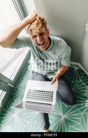 Young man sitting on ground, holding laptop, screaming in despair Stock Photo