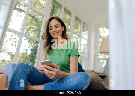 Smilong mature woman sitting on couch at home using cell phone with man in background Stock Photo