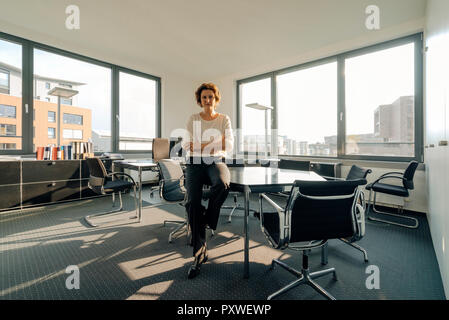 Successful businesswoman sitting on desk in her office with arms crossed Stock Photo