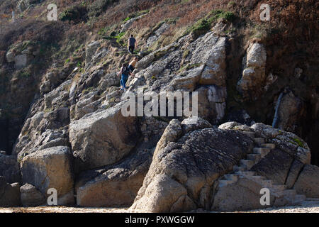 Visitors to Porthcurno beach climbing the steep, rocky steps and path towards the Minnack Theatre above on the Cornish coastal path. Stock Photo