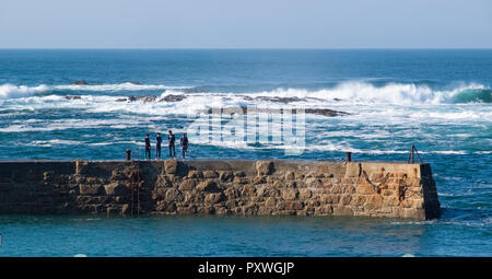 Distant view of four men in wet suits standing on Sennen Cover harbour wall looking out to sea at the massive breaking waves over rocks Stock Photo