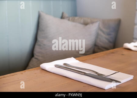 Simple place setting in a restaurant of cutlery on a white napkin and pale wood table with two grey cushions and pale blue walls Stock Photo