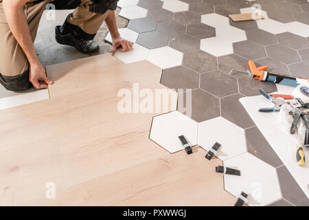 Male worker installing new wooden laminate flooring. The combination of wood panels of laminate and ceramic tiles in the form of honeycomb. Kitchen renovation. Stock Photo