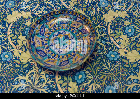 A brightly coloured and multi patterned asian style plate in turquoise, blues and gold hung on a wall covered in wallpaper of similar patterns Stock Photo