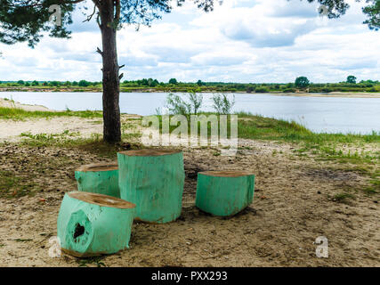 Unusual chairs and a table made of a round tree trunk painted green on the bank of the river against the backdrop of a tree and a blue sky with white  Stock Photo