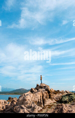 a young caucasian man seen from behind, on the top of a rock formation, facing the Mediterranean sea in the Southern coast of Corsica, in France Stock Photo
