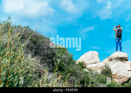 a young caucasian man, seen from behind, on the top of a rock formation taking a photo of the landscape in the Sout of Corsica, in France, with his sm Stock Photo