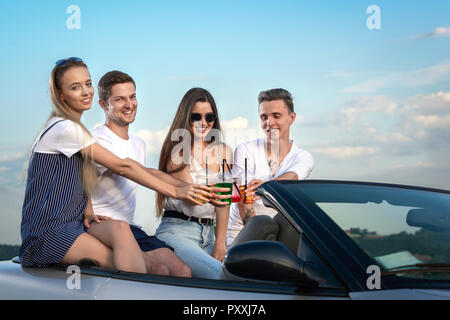 Company of four friends sitting together on cabriolet and clinking glasses with lemonade. Pretty girls and young handsome men having fun and enjoying summertime adventures. Stock Photo