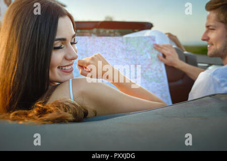 Charming, gorgeous young woman turning to camera while sitting with husband in cabriolet. Adorable model having long eyelashes, smooth skin and chestnut long hair. Man holding map an looking away. Stock Photo