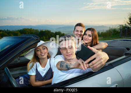 Two beautiful couple sitting in cabriolet and making selfie. Handsome boy holding phone and friends looking at gadget. Company spending summertime together, traveling by car. Stock Photo