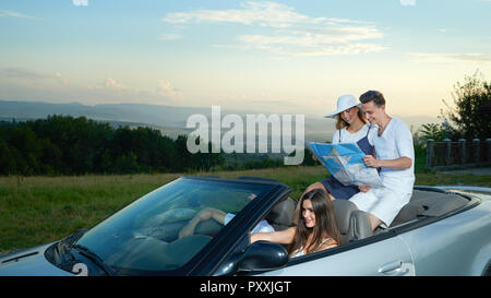 Company traveling by car, spending summertime together. Couple sitting on silver cabriolet, holding map and planning route. Young brunette driving car, another girl wearing in white brim broad hat. Stock Photo