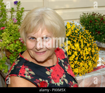 Close-up head and shoulders portrait of smiling senior blonde woman in shift dress on bright flowers background. Stock Photo