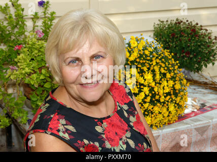 Close-up head and shoulders portrait of smiling senior blonde woman in shift dress on bright flowers background. Stock Photo