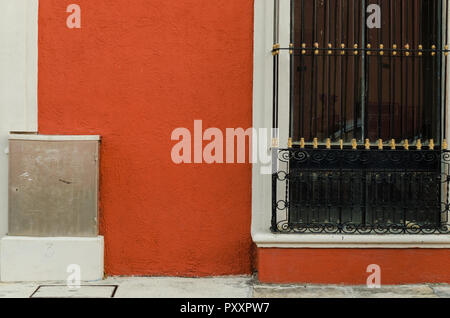 Window with black bars and a metal box on one side on an orange wall, side view on the sidewalk of an urban street. Stock Photo
