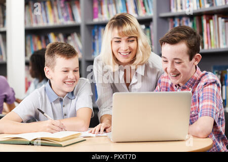 Female Teacher Helping Two Male High School Students Working At Laptop In Library Stock Photo