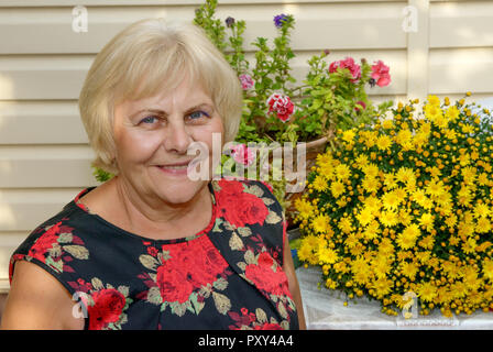 Close-up head and shoulders portrait of smiling senior blonde woman in shift dress on bright flowers background. Stock Photo