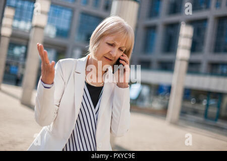 Outdoor portrait of angry senior businesswoman who is talking on phone. Stock Photo