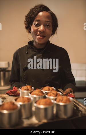Smiling baker carrying a tray of muffins in her kitchen Stock Photo