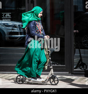 Muslim woman in hijab riding kick scooter. Muslim woman wearing headscarf riding an adult push scooter on city pavement Stock Photo