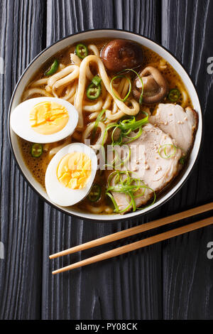 Asian style soup with udon noodles, pork, boiled eggs, mushrooms and green onions close-up in a bowl on the table. Vertical top view from above Stock Photo