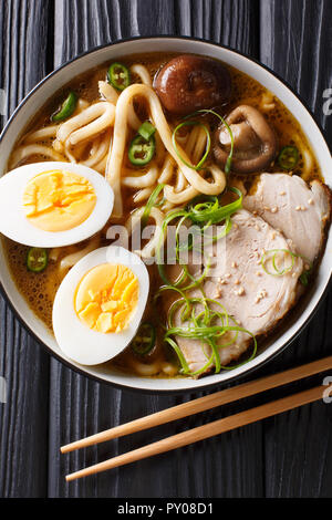 Spicy udon noodle soup, pork, boiled eggs, shiitake and onions close-up in a bowl on the table. Vertical top view from above Stock Photo