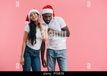 concentrated african american man in chrismas hat eavesdropping girlfriend talking on smartphone isolated on pink background Stock Photo