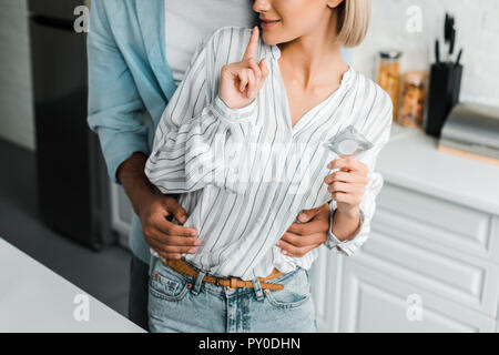 cropped image of girlfriend showing silence gesture and holding condom in kitchen Stock Photo