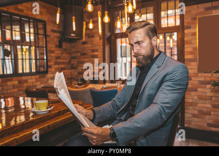 Portrait of a young bearded businessman sitting at the bar, reading newspapers and drinking coffee. Stock Photo