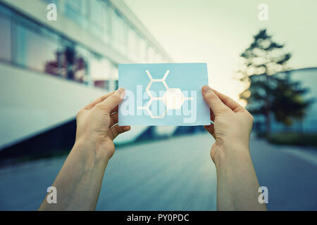 Close up of woman hands holding a paper sheet with scientific cell structure symbol inside, over laboratory building background. DNA molecule grid for Stock Photo