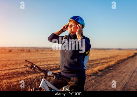 Middle-aged woman bicyclist taking on and fastens helmet in autumn field at sunset. Senior sportswoman enjoying hobby. Stock Photo