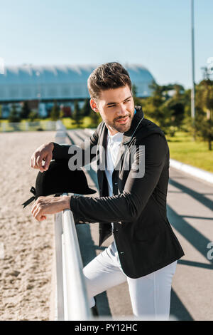 handsome male equestrian leaning on fence with riding helmet at horse club Stock Photo