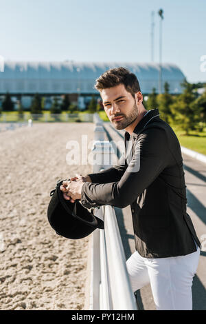 handsome equestrian in white breeches and black jacket leaning on fence with riding helmet at horse club Stock Photo