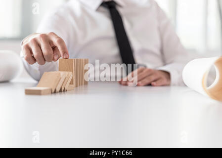 Businessman wearing white shirt and black necktie interrupting domino effect by stopping wooden dominoes bricks from crumbling. Stock Photo
