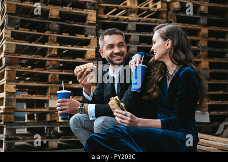 side view of smiling couple in luxury clothing with hot dogs and soda drinks sitting on wooden pallet on street Stock Photo