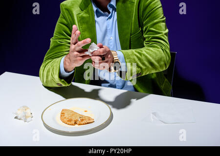 cropped shot of man in velvet jacket drying hands with napkin while sitting at table with meat cheburek on plate with blue background Stock Photo
