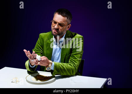 man in stylish green velvet jacket drying hands with napkin at table with meat pastry on plate with blue background behind Stock Photo