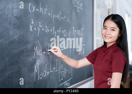 Beautiful Asian student writing on blackboard with chalk in classroom. Stock Photo