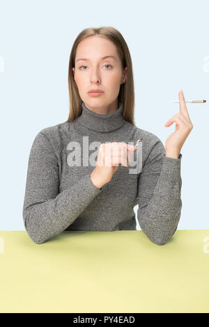 The young woman smoking cigarette while sitting at table at studio. Trendy colors. The portrait of caucasian girl in minimalism style with copy space Stock Photo