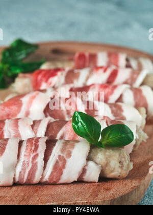 Cold prepared to grill pigs sausages wrapped in bacon on plate. Raw sausages wrapped in bacon served fresh green basil leaves on wooden cutting board. Shallow DOF. Copy space for text. Stock Photo