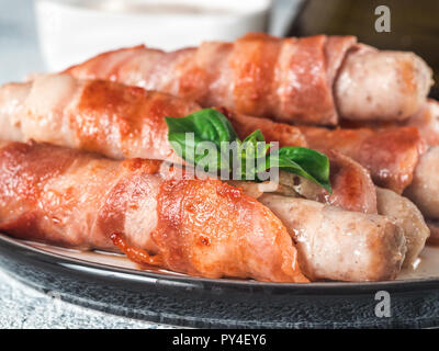 Ready-to-eat pigs sausages wrapped in bacon on plate. Fried savory sausages wrapped in bacon served fresh green basil leaves with sauce on background. Shallow DOF. Copy space for text. Stock Photo