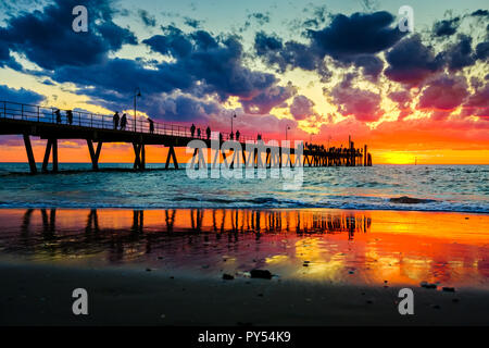 People stroll on Glenelg jetty at sunset sky splashed with colourful hues of red, orange and blue in Adelaide, South Australia Stock Photo