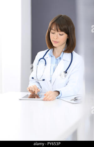 Doctor typing on laptop computer while sitting at the white table in hospital office. Physician at work. Medicine and healthcare concept. Stock Photo