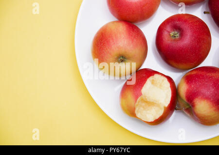 several whole red apples and one bitten in a plate on a yellow background of autumn composition and concept. Stock Photo