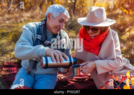 Senior couple having tea in autumn forest. Happy man and woman enjoying picnic and nature. Smiling man pouring tea out of thermos Stock Photo