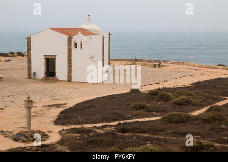 Landscape with a sandy land and Atlantic ocean.  White church with bells and a cross. Cloudy sky. Fortress in Sagres, Portugal. Stock Photo