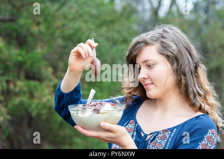 Young woman outside, outdoors, holding glass bowl of homemade, raw vegan vanilla ice cream, adding chocolate liquid sauce from spoon, looking down Stock Photo