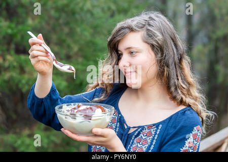 Young woman outside, outdoors, holding glass bowl of homemade, raw vegan vanilla ice cream with chocolate liquid sauce, holding spoon, ready to eat Stock Photo