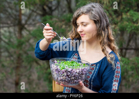 Young woman outside, outdoors, holding glass bowl of homemade red, purple cabbage salad dish with green onions, scallions with spoon in hand, eating Stock Photo