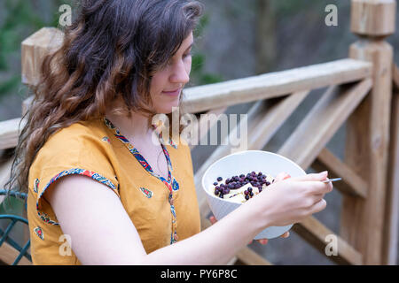 Closeup of young woman, female sitting outside on wooden home, house deck, holding eating raw vegan fruit salad from bilberry, banana, apples, blueber Stock Photo